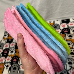 May include: A stack of three soft, plush cloths in pink, light blue, and lime green. The cloths are folded and held in a hand.