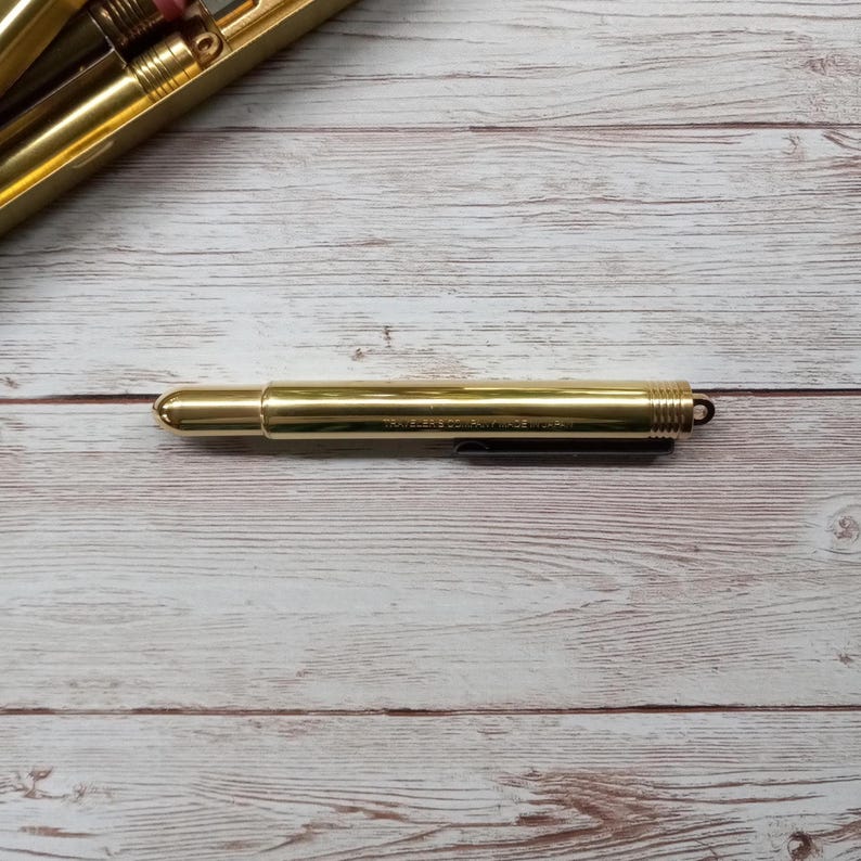 May include: A gold-coloured pen with a rounded tip and a black clip. The pen is lying on a light-coloured wooden surface. The pen has the text "TRAVELER'S COMPANY MADE IN JAPAN" printed on the side.