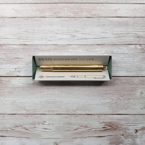 May include: A gold-coloured brass fountain pen in its packaging. The pen is lying horizontally inside a green and white box. The box has the words "BRASS FOUNTAIN PEN" printed on it. The pen is a shiny gold colour.