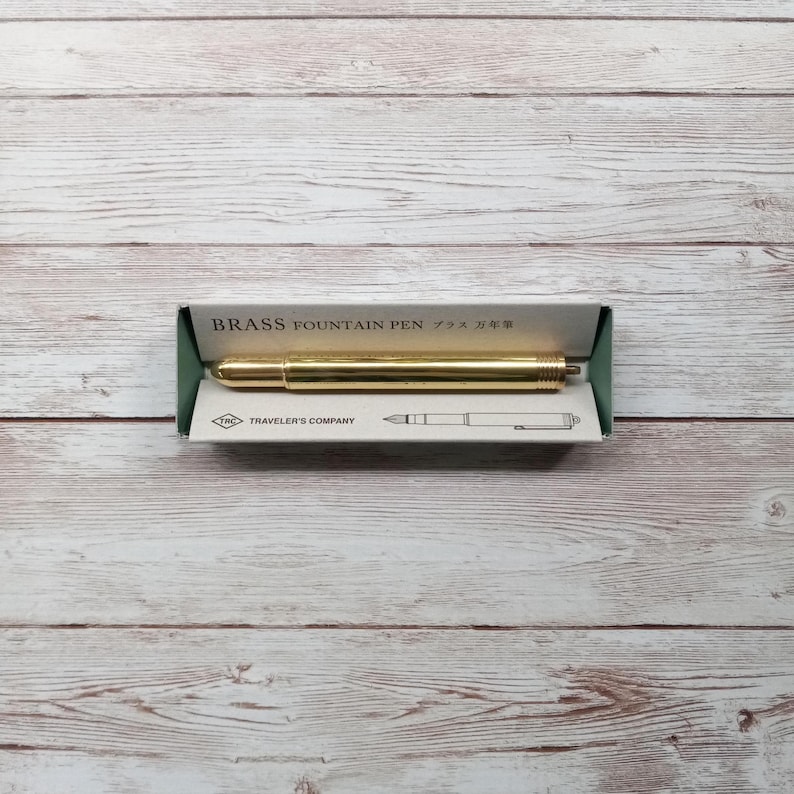 May include: A gold-coloured brass fountain pen in its packaging. The pen is lying horizontally inside a green and white box. The box has the words "BRASS FOUNTAIN PEN" printed on it. The pen is a shiny gold colour.