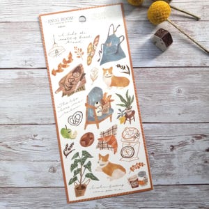 May include: A sticker sheet with a cozy bread theme. Illustrations include a corgi, bread, an apron, a chair, and plants. The text "Snug Room" and "I like the smell of fresh bread" are also present. The color palette is soft and warm.