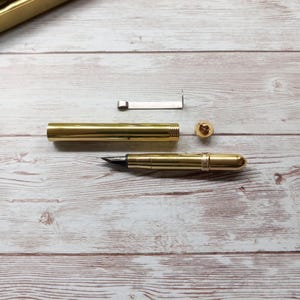 May include: A disassembled gold fountain pen with a black nib, cap, and other parts. The pen is on a light-coloured wooden surface. The pen's components are separated, revealing the internal parts.