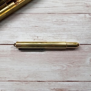 May include: A gold-coloured pen with a cylindrical body and a rounded tip. The pen has a small loop at one end and a black clip. The pen is lying on a light-coloured wooden surface.