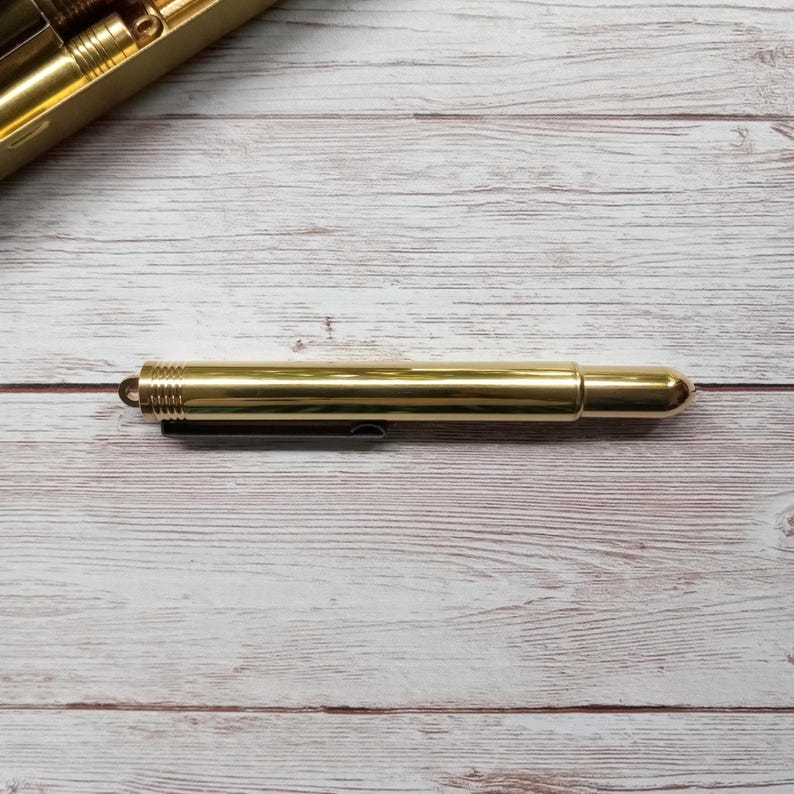 May include: A gold-coloured pen with a cylindrical body and a rounded tip. The pen has a small loop at one end and a black clip. The pen is lying on a light-coloured wooden surface.