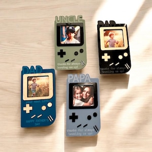 May include: Four picture frames shaped like retro game consoles. The frames are in shades of blue, green, and black, each with a photo inside. Text includes "UNCLE", "DAD", and "PAPA", along with phrases like "thanks for always leveling me up!" and "HAPPY FATHER'S DAY".