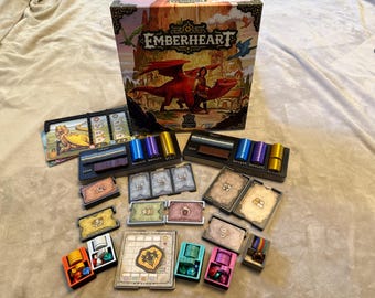 Pode incluir: Caixa do jogo de tabuleiro Emberheart com uma ilustração de dragão, peças do jogo, cartas e fichas de jogador. A caixa apresenta o título e a arte do jogo. Vários componentes do jogo estão dispostos ao redor da caixa, incluindo cilindros coloridos e cartas.