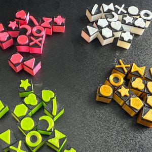 Puede incluir: Un surtido de piezas de juego coloridas y de forma geométrica. Las piezas son de varios colores, incluyendo rosa, blanco, verde y amarillo, con acentos negros. Las formas incluyen corazones, estrellas y círculos. Las piezas están dispuestas sobre una superficie oscura.