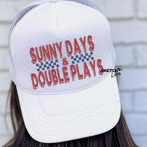 May include: White trucker cap with a mesh back and a curved peak. The cap features the text "SUNNY DAYS & DOUBLE PLAYS" in red and blue lettering with a checkered design.
