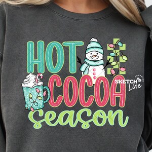 May include: Dark gray sweatshirt with the words "HOT COCOA SEASON" in green and red, along with a graphic of a snowman, a cup of cocoa, and string lights. The sweatshirt is worn by a person in blue jeans.