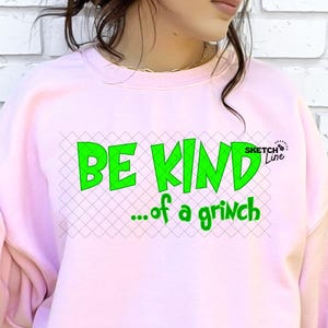 Op de afbeelding: Een lichtroze sweatshirt met de tekst "BE KIND...of a grinch" in het groen. Het sweatshirt wordt gecombineerd met een donkerblauwe jeans. Een rode beker met een ruitpatroon wordt in de linkerhand gehouden.