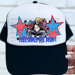 Può includere: Un cappello da camionista nero e bianco con un pannello frontale bianco. Il pannello frontale presenta un'immagine di un cartone animato di una persona con un abito rosso, bianco e blu seduta su un trono con due stelle rosse dietro di lei. Il testo "HERE COMES THE BOOM" è stampato sotto l'immagine.