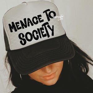 Può includere: Cappellino da camionista bianco e nero con la scritta "MENACE TO SOCIETY" in grassetto nero. Il cappello ha un retro in rete e una visiera curva. La persona indossa anche una maglietta nera.