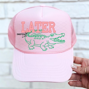 May include: Pink trucker hat with the word "LATER" in coral letters and a green alligator graphic. The hat has a mesh back and a curved brim. The text "Sketch Line" is on the left side of the hat.