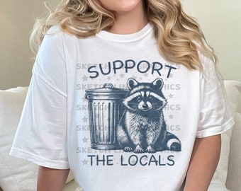 May include: A white t-shirt with a blue graphic of a raccoon in a trash can. The text "SUPPORT THE LOCALS" is printed below the graphic.