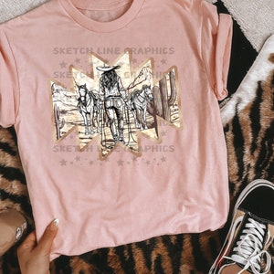 Może przedstawiać: R&oacute;żowy T-shirt z złotym nadrukiem przedstawiającym kobietę jeżdżącą konno w pustynnym krajobrazie. Nadruk zawiera tekst "Sketch Line Graphics".