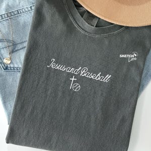 May include: A casual fashion ensemble featuring a gray t-shirt with the text "Jesus and Baseball" and a baseball graphic, light-wash denim jeans, and a tan fedora hat. A gold necklace and dried flowers complete the look.