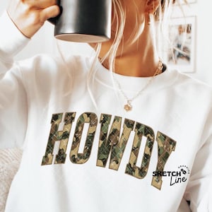 May include: White sweatshirt with the word "HOWDY" in camouflage print across the chest. The sweatshirt also has the text "Sketch Line" in a small font on the lower right side. A black mug is held up to the face.