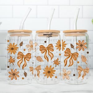 Op de afbeelding: Drie doorzichtige glazen bekers met houten deksels en glazen rietjes. Elke beker is versierd met herfstthema-afbeeldingen, waaronder oranje strikken, bloemen en bladeren. De bekers zijn ongeveer 15 cm hoog.