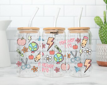 May include: Three clear glass tumblers with bamboo lids and reusable straws. Each tumbler features a different design with colorful illustrations of school supplies, including apples, pencils, scissors, and a globe. The text "teacher life" and "teacher life" is featured on two of the tumblers.