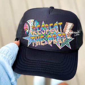 May include: Black trucker hat with a baseball graphic and the text "RESPECT THE DRIP" in colorful letters. The hat has a mesh back and a curved brim. The baseball graphic includes a baseball and a baseball bat.