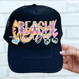 May include: Black trucker hat with the words "Beach Vibes" in a colorful, tropical design. The graphic includes a surfboard, flowers, hearts, and palm leaves. The hat has a mesh back and a curved brim.