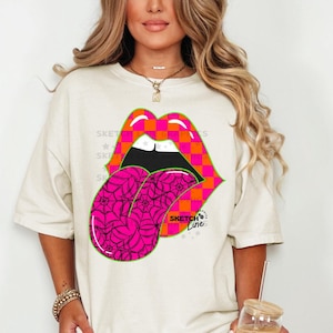 May include: Cream-colored t-shirt featuring a graphic of stylized lips with a tongue sticking out. The lips are orange and pink checkered, outlined in green, and the tongue is bright pink with a spiderweb design. The text "SKETCH Line" is visible.