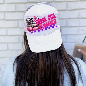 Puede incluir: Gorra de camionero blanca con parte trasera de malla y visera curva. La parte delantera presenta un gr&aacute;fico de un mapache con gafas de sol y sombrero, tumbado en un flotador de flamenco rosa. El texto "FERAL GIRL SUMMER" est&aacute; en rosa y morado.