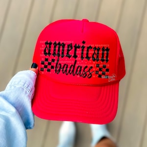May include: A vibrant red trucker hat with the words "American badass" in black script and gothic lettering. The hat features a mesh back and a curved brim, perfect for sun protection. The hat is a bold fashion statement.
