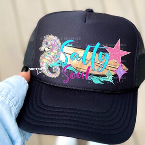 Peut inclure: Casquette de camionneur noire avec un graphique coloré de cheval de mer, de planche de surf et d'étoiles. Le texte "Salty Soul" est écrit sur la planche de surf.