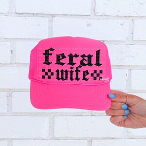May include: A bright pink trucker hat with the words "feral wife" in black gothic lettering. The hat has a mesh back and a curved brim. The text is centered on the front of the hat.