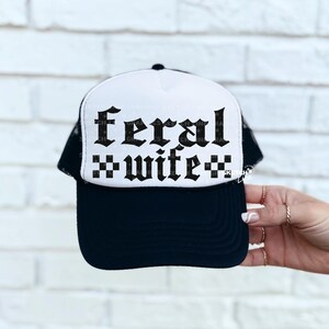 May include: Black and white trucker hat with the words "feral wife" in a gothic font. The hat has a white front panel with the text and a black mesh back. The brim is black.
