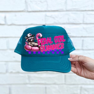 Puede incluir: Gorra de camionero turquesa con un dise&ntilde;o de un mapache con gafas de sol en un flotador de flamenco rosa. El texto "FERAL GIRL SUMMER" es rosa y morado. Un divertido accesorio de verano.