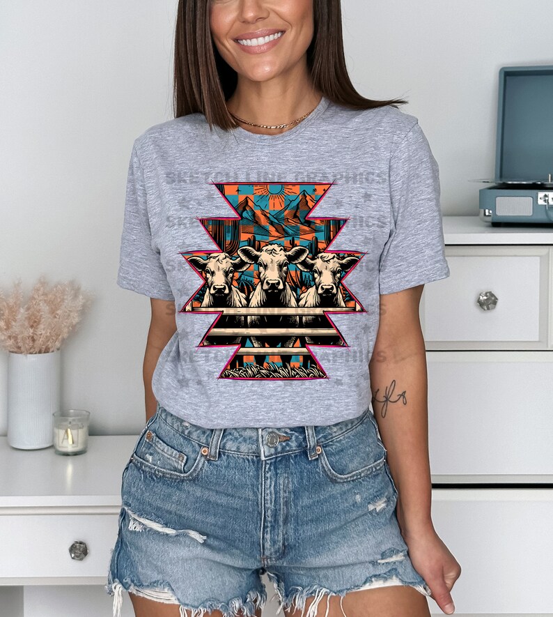 May include: A gray t-shirt with a graphic of three cows in front of a fence with a mountain and sunset scene behind them. The graphic is in a geometric pattern with pink, orange, blue, and green colors.