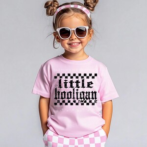 K&ouml;nnte beinhalten: Ein pinkfarbenes T-Shirt mit einem schwarzen Karomuster und dem Text "little hooligan" in gotischer Schrift.