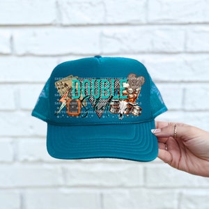 May include: Teal blue trucker hat with a mesh back. The front of the hat features a colorful graphic with the text "DOUBLE DOWN" and playing cards, dice, and a lightning bolt.
