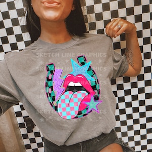 Może przedstawiać: Szary T-shirt z graficznym wzorem przedstawiającym turkusową podkowę z wystającym językiem w różowo-niebieską kratkę, fioletowym błyskawicą i turkusową gwiazdą z brokatem. Na koszulce nadrukowany jest tekst "Sketch Line Graphics".