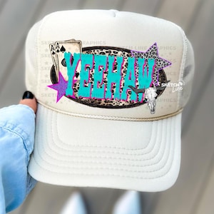 Puede incluir: Una gorra de camionero blanca con una malla en la parte posterior. La parte delantera de la gorra presenta un gráfico colorido con la palabra "Yeehaw" en letras turquesa, rosa y negras. El gráfico también incluye un diseño de estampado de leopardo, cartas de juego, una estrella y una calavera.