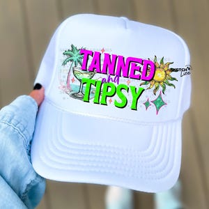 Puede incluir: Gorra de camionero blanca con un diseño gráfico colorido que dice "Tanned and Tipsy". El diseño incluye una palmera, una copa de cóctel, un sol y estrellas.
