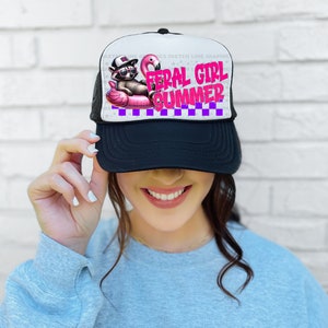 Puede incluir: Gorra de camionero en blanco y negro con el texto "FERAL GIRL SUMMER" en rosa y morado. La gorra presenta un gr&aacute;fico de un mapache con gafas de sol y sombrero, flotando en un flotador de flamenco rosa.