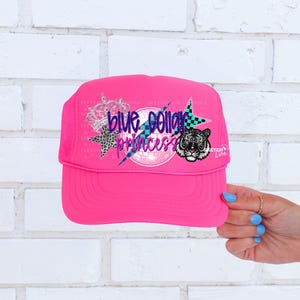 May include: Pink trucker hat with a blue, purple, and white graphic that says "Blue Collar Princess" with a cheetah print star, a checkered pattern, and a tiger face.