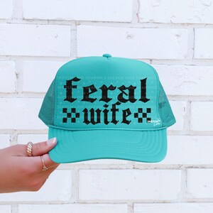 May include: A turquoise trucker hat with a mesh back and a curved brim. The front of the hat features the words "feral wife" in a gothic font. The hat is set against a white brick wall.