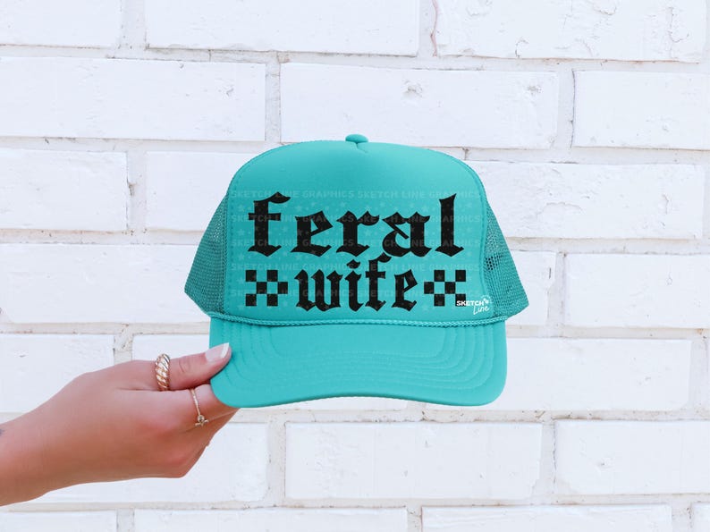 May include: A turquoise trucker hat with a mesh back and a curved brim. The front of the hat features the words "feral wife" in a gothic font. The hat is set against a white brick wall.
