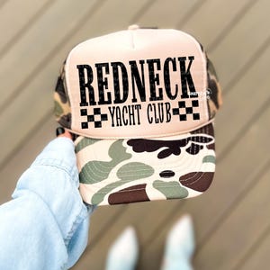 Puede incluir: Gorra de camionero con un panel frontal beige y un estampado de camuflaje en la visera y los laterales. La gorra presenta el texto "REDNECK YACHT CLUB" en letras negras.