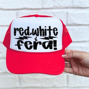May include: A red and white trucker hat with the words "red.white & feral" in black script. The hat has a red brim and mesh back, with a white front panel. The text is centered on the front panel.