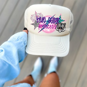 May include: A white trucker hat with a blue, pink, and black design that says "Blue Collar Princess". The design also includes a crown, a star, and a tiger.