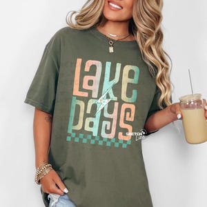 May include: A green t-shirt with a graphic design that says "lake days" in a colourful, layered font with a lightning bolt graphic. The shirt has a relaxed fit and short sleeves.