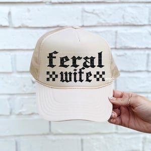 May include: A cream-colored trucker hat with a mesh back and a curved brim. The front of the hat features the words "feral wife" in a gothic font. The hat is held by a hand with multiple rings.