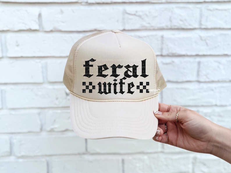 May include: A cream-colored trucker hat with a mesh back and a curved brim. The front of the hat features the words "feral wife" in a gothic font. The hat is held by a hand with multiple rings.