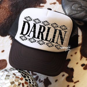 May include: A trucker hat with a white front panel and black mesh back. The front features the word "DARLIN" in bold black letters, with a geometric design above and below. The brim is brown.