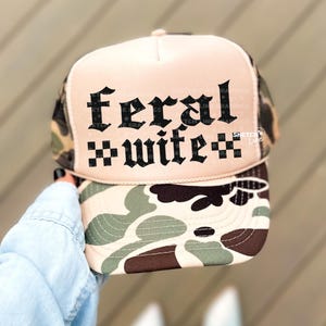 以下が含まれることがあります： ピンク色のフロントパネルと迷彩柄のつばが付いたトラッカーハット。帽子には黒いゴシック体で「feral wife」の文字が書かれています。メッシュの背面はタン色です。薄いブルーのデニムシャツを着た人が帽子を持っています。